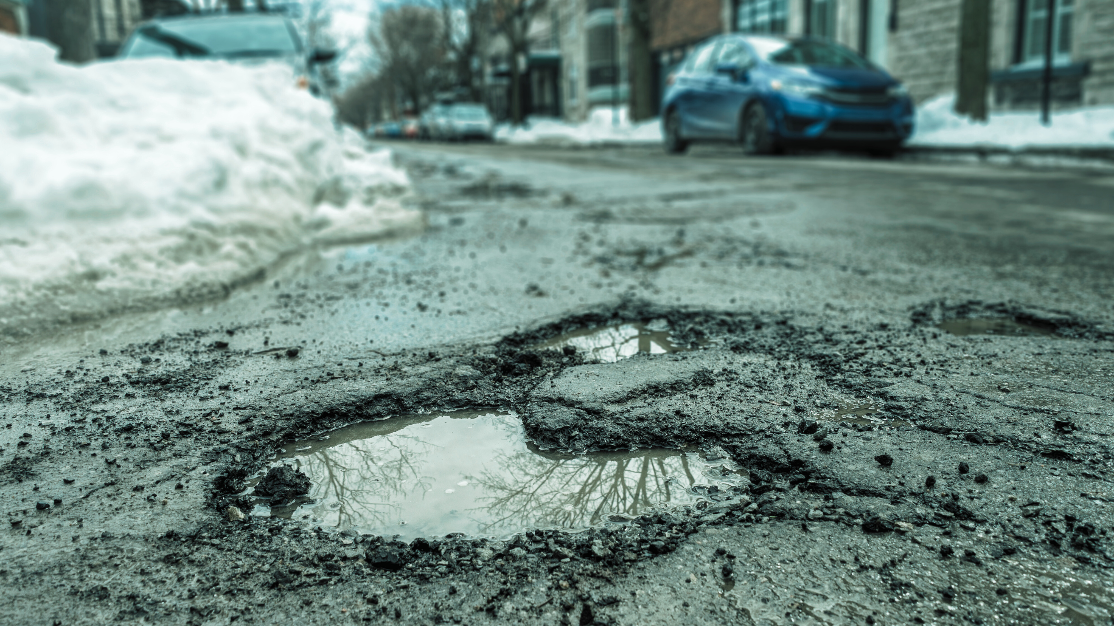 In the foreground, a large pothole in an urban street, in the background piles of snow and cars parked.