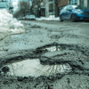 In the foreground, a large pothole in an urban street, in the background piles of snow and cars parked.